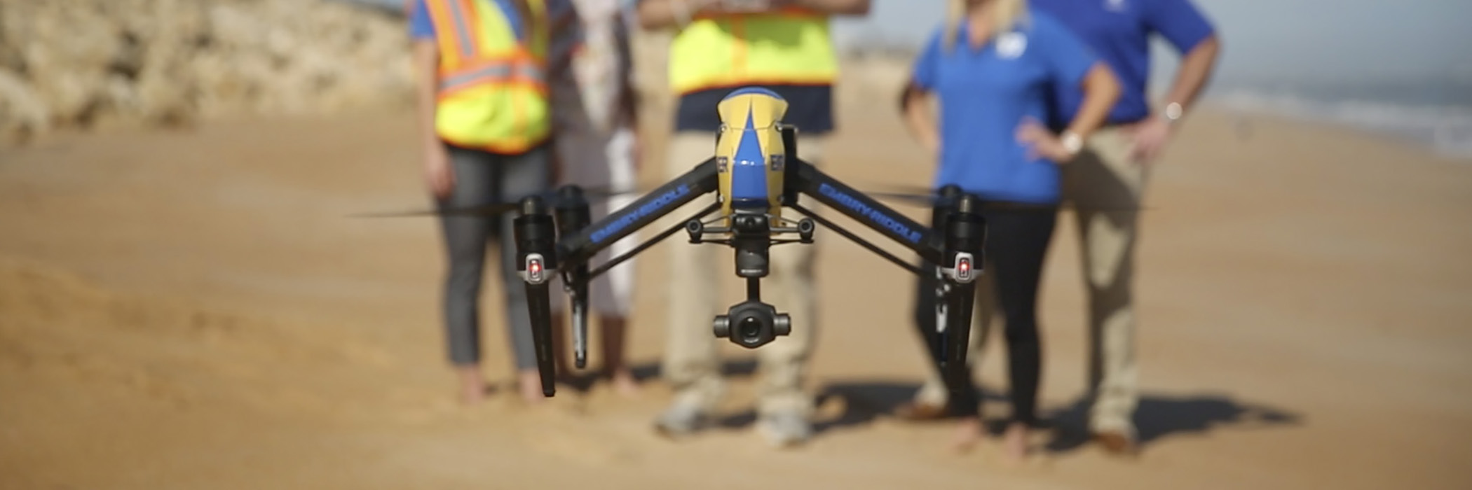 Drone hovering low over beach with operating crew in background